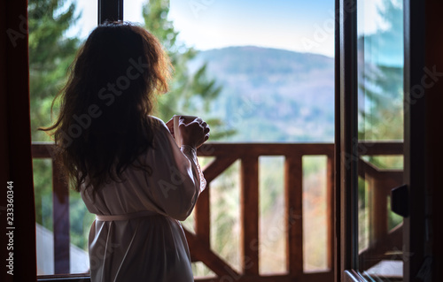 Woman in the morning holding a cup of tea or coffee and looking at the mountains standing near the window .
