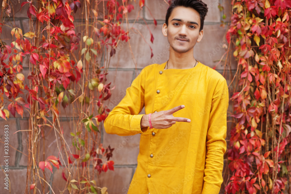 Indian stylish man in yellow traditional clothes posed outdoor against red leaves wall, shows two fingers. Indian stylish man in yellow traditional clothes posed outdoor against red leaves wall, shows two fingers.