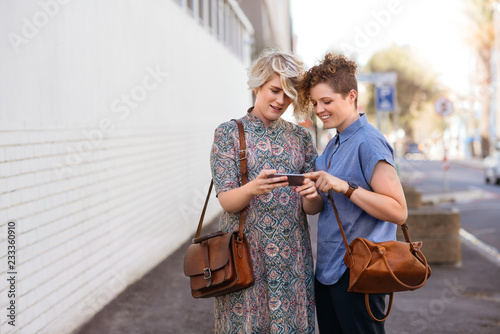 Smiling lesbian couple standing outside looking at cellphone photos