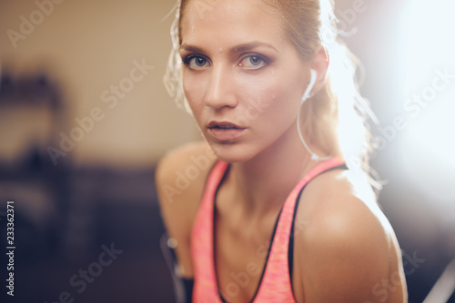 Close up of woman looking at camera. Sweat on face, earphones in ears, back light. Gym interior, healthy lifestyle concept.