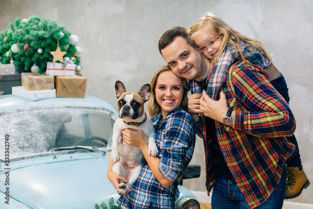 Beautiful family in checked shirts standing with dog near blue retro car with presents on the roof. Family in stylish decorated interior studio for Christmas and New Year holidays Beautiful family in checked shirts standing with dog near blue retro car with presents on the roof. Family in stylish decorated interior studio for Christmas and New Year holidays