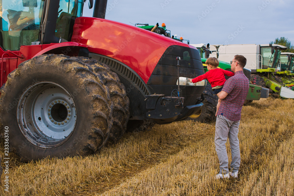 The happy father standing in the field near the tractors and holding his little son in arms The happy father standing in the field near the tractors and holding his little son in arms