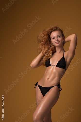 Young sexy slim tanned woman in black swimsuit posing against yellow background. Fashion portrait of beautiful girl with curly hair. Swimwear or bikini model
