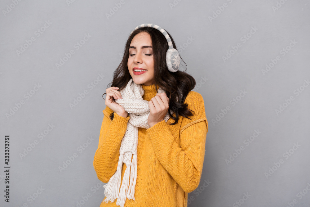 Portrait of cheerful woman wearing ear muffs and scarf smiling and touching scarf, isolated over gray background Portrait of cheerful woman wearing ear muffs and scarf smiling and touching scarf, isolated over gray background
