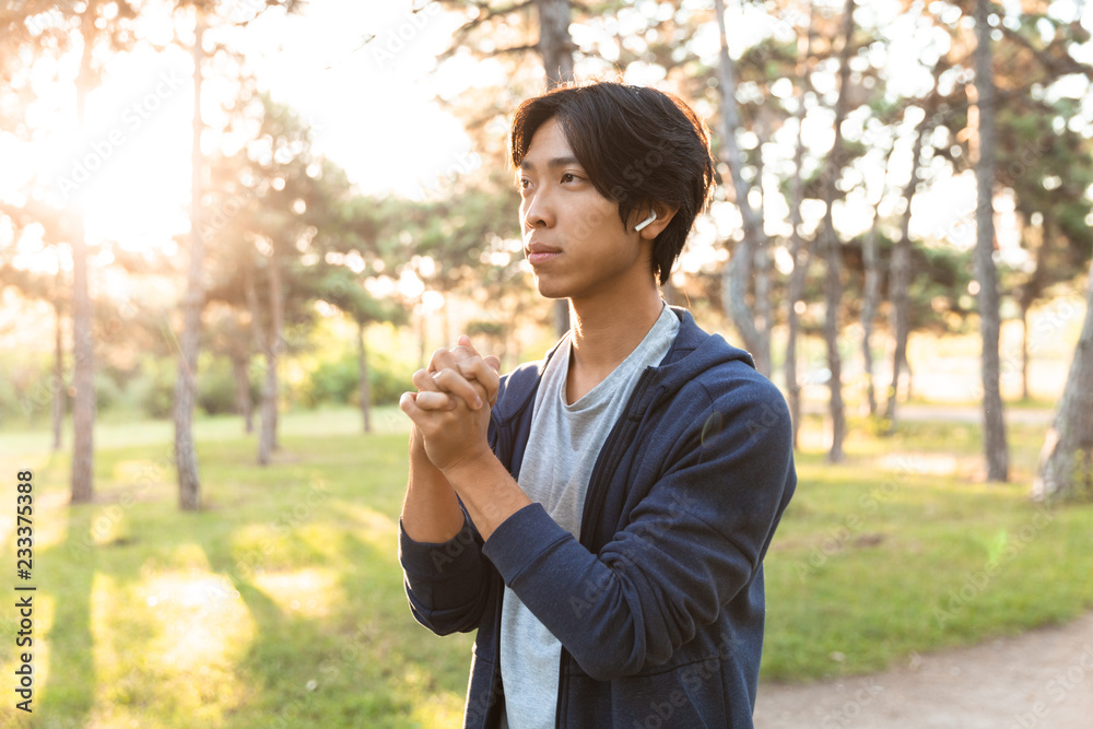 Image of sporty asian guy in casual wear and eyeglasses, working out and stretching his body in park with green trees Image of sporty asian guy in casual wear and eyeglasses, working out and stretching his body in park with green trees