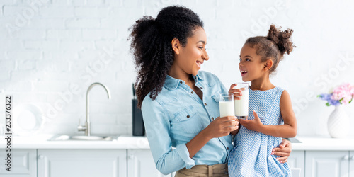 фотографія smiling african american mother and daughter holding glasses of milk in kitchen