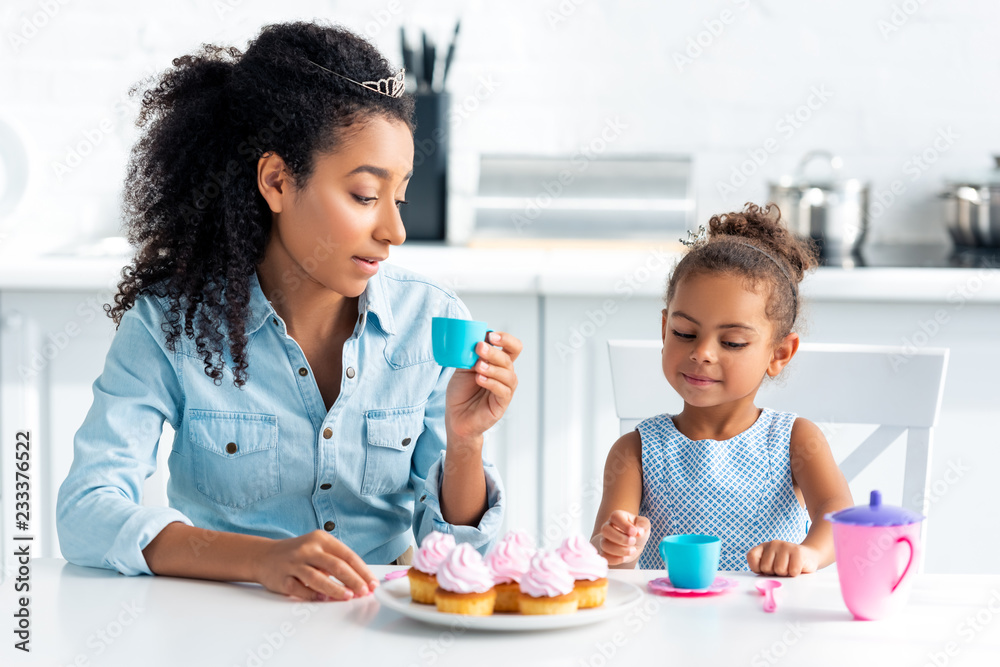 african american mother and daughter with tiaras sitting at table with cupcakes in kitchen african american mother and daughter with tiaras sitting at table with cupcakes in kitchen