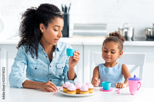 african american mother and daughter with tiaras sitting at table with cupcakes in kitchen