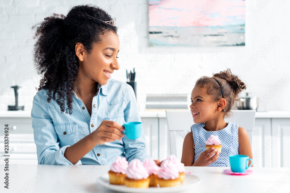 smiling african american mother and daughter with tiaras holding plastic cup and cupcake in kitchen smiling african american mother and daughter with tiaras holding plastic cup and cupcake in kitchen