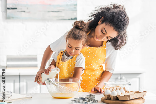 African American mother and daughter preparing dough and pouring milk into bowl in kitchen photo