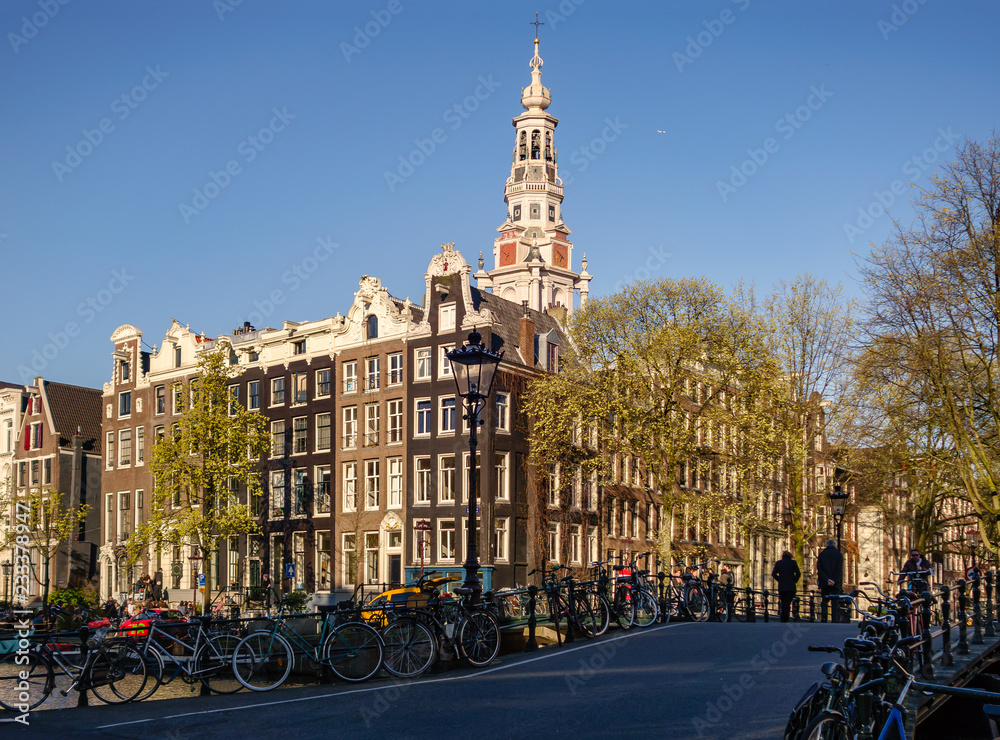 Obraz premium Gingerbread houses near the canal, a bridge with ancient lanterns, indispensable bicycles, idle Amsterdamers staggering under the rays of a still hot sun, a southern church and an airplane in the sky.