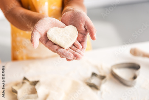 cropped image of african american kid holding heart shaped unbaked cookie in hands in kitchen photo