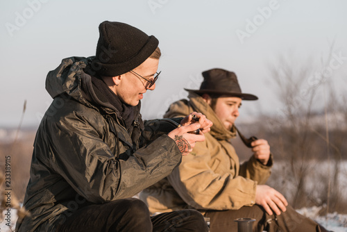Men is smoking tobacco-pipe and drink coffee. Outdoor portrait of hikker in black sunglasses sitting outdoors during sunny winter day.