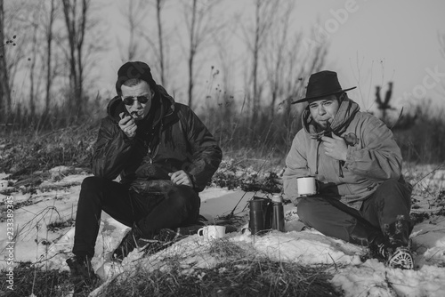Men is smoking tobacco-pipe and drink coffee. Outdoor portrait of hikker in black sunglasses sitting outdoors during sunny winter day. photo