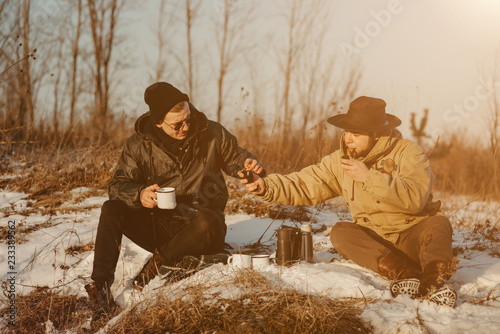 Men is smoking tobacco-pipe and drink coffee. Outdoor portrait of hikker in black sunglasses sitting outdoors during sunny winter day. © vitleo