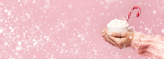 Female hands in knitted sweater holding cup of marshmallows and Christmas candy cane on pink background Flat Lay copy space. Winter traditional food Festive decor celebration presents Xmas holiday