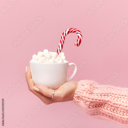 Female hands in knitted sweater holding cup of marshmallows and Christmas candy cane on pink background Flat Lay copy space. Winter traditional food Festive decor celebration presents Xmas holiday