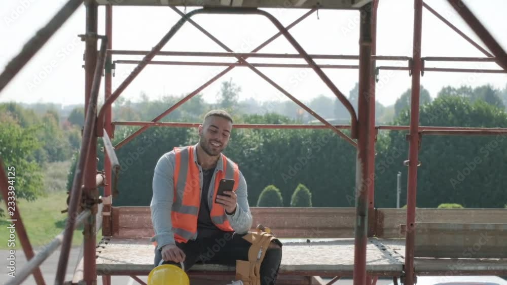 Person working in construction site. White young man at work in new house inside apartment building. Latino manual worker smiling and using smartphone for internet and social media during break