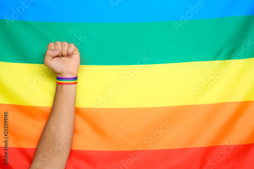 Man wearing gay wristband on hand near rainbow flag  closeup. Space for text