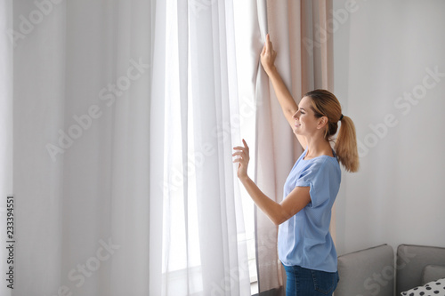 Woman opening curtains and looking out of window at home. Space for text