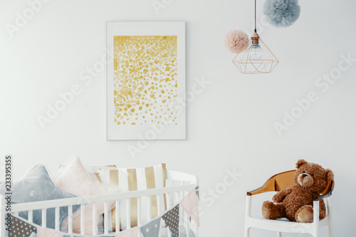Abstract golden painting in white frame above wooden crib with pillows and chair with teddy bear in scandinavian baby room interior