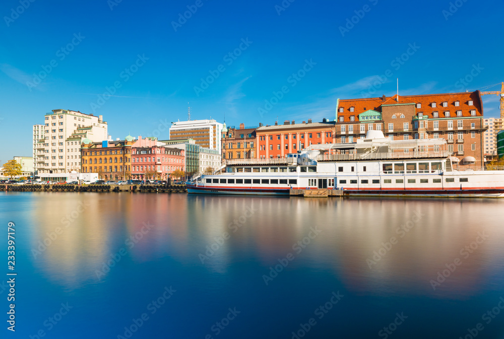 Skyline of a city reflected in water. Cityscape of Malmo with historic and modern architecture, embankment and ship, long exposure photo. Oresund region, Sweden