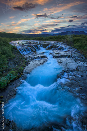The waterfall Bruarfoss with golden clouds in the sky. The flowing water is captured by a long exposure. Amazing blue color of water from the glacier. Natural and colorful environment.