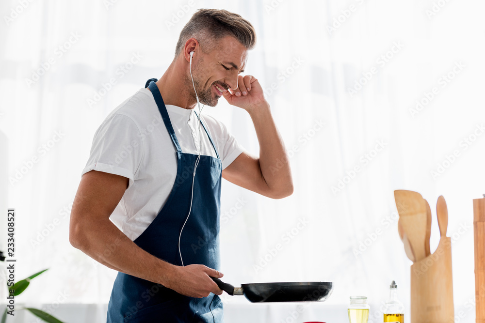 smiling man with earphones and pan in hand listening to music at kitchen