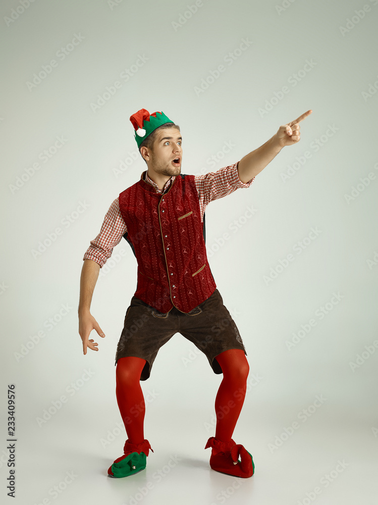 The happy smiling friendly man dressed like a funny gnome or elf pointing to left on an isolated gray studio background. The winter, holiday, christmas concept