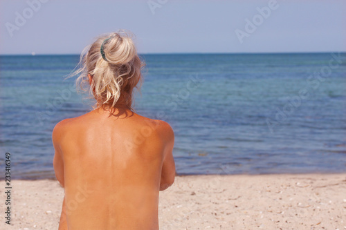 Beautiful blond sun tanned topless woman sitting on beach in sunshine and looking over the sea - seen from behind with copy space © Ole