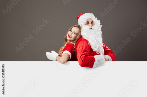 Santa Claus and young mrs. Claus stand back to back  behind a white canvas and leans on it on the gray background.