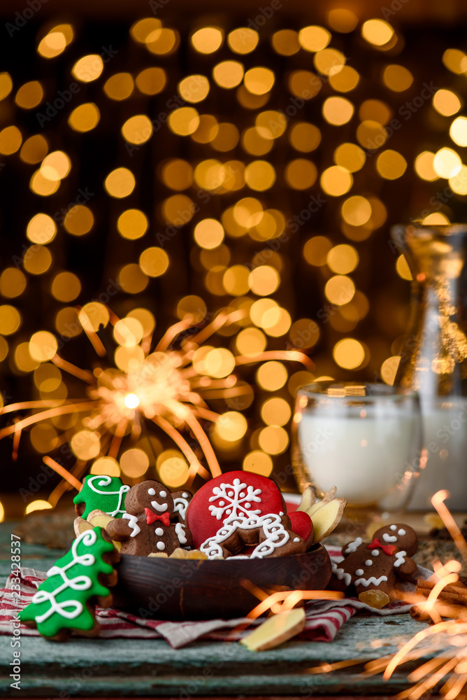 Obraz premium Gingerbread mittens, little man, snowflakes, christmas trees in a brown wooden plate and a bottle of milk on a green wooden on the background of blurred garland. close up. space