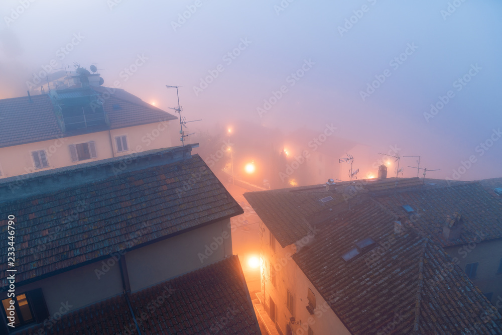 High angle, aerial view on foggy misty weather, fog, mist in morning with houses, buildings, roofs, rooftops, street lights, lamps in Chiusi, Italy, Umbria