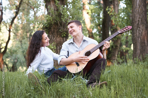 young couple sitting in the forest and playing guitar, summer nature, bright sunlight, shadows and green leaves, romantic feelings