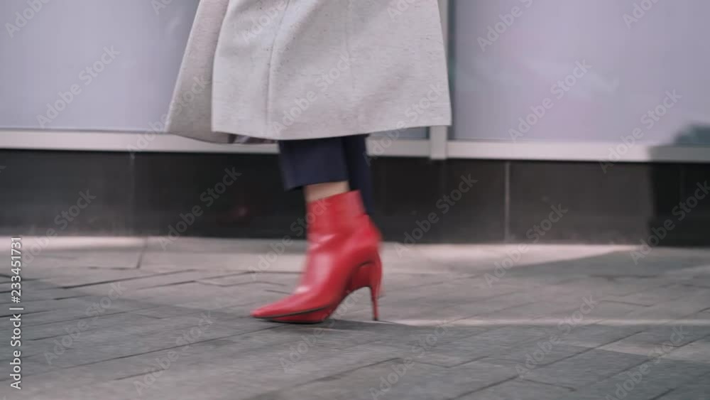 Legs of unrecognizable woman wearing black pants, white coat and red boots walking in autumn street. Tracking slow motion medium shot Legs of unrecognizable woman wearing black pants, white coat and red boots walking in autumn street. Tracking slow motion medium shot