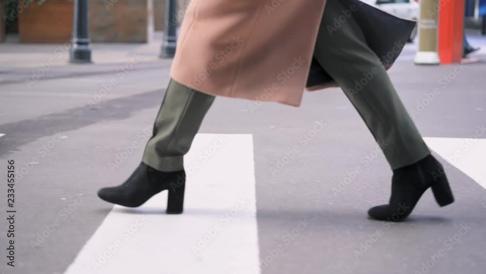 Legs of unrecognizable woman wearing beige coat, khaki pants and black boots crossing the road. Tracking slow motion medium shot Legs of unrecognizable woman wearing beige coat, khaki pants and black boots crossing the road. Tracking slow motion medium shot