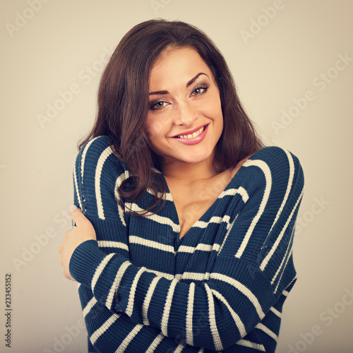 Happy woman hugging herself with natural emotional enjoying face in warm winter sweater on blue background. Love concept of yourself. Toned vintage portrait