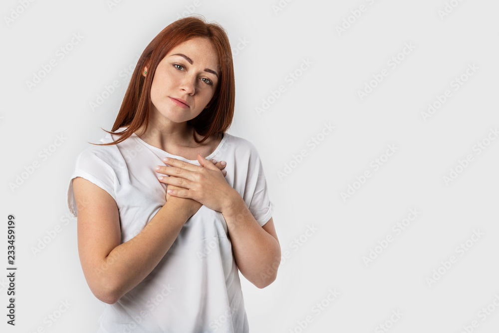 Portrait of a red-haired young woman keeps hands on chest, expresses sympathy. Kind hearted friendly nice girl shows kindness, wearing blank white t-shirt Portrait of a red-haired young woman keeps hands on chest, expresses sympathy. Kind hearted friendly nice girl shows kindness, wearing blank white t-shirt
