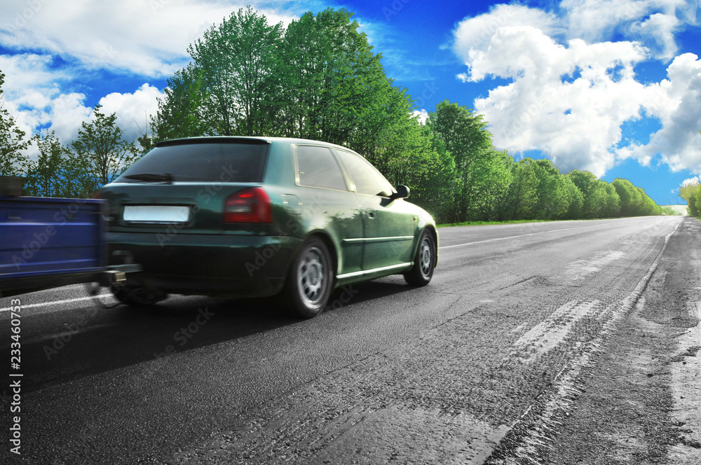 Fototapeta premium A green car with a trailer on the countryside road against sky with clouds