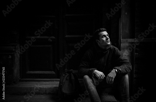 A young man with a hipster hanging on an outer garment on the stairs. Hoodshavy man in a wool jacket and high quality slippers with a backpack. Conceptual black and white photo.