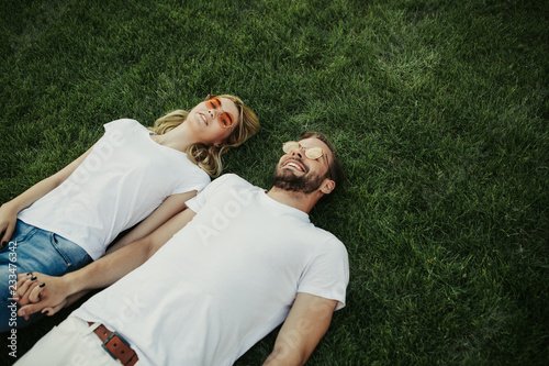 Top view of blithesome young twosome is lying on green lawn and holding hands. They wearing sunglasses. Copy space on right side