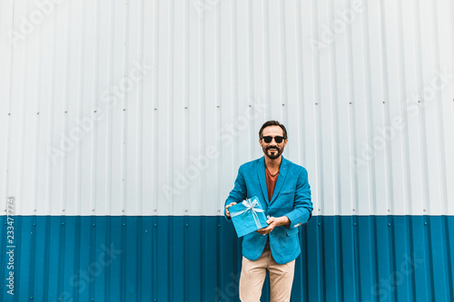 Lovely present. Cheerful bearded young man in stylish clothes standing near the wall and smiling while showing a beautiful blue present