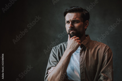 Portrait of pensive unshaven male keeping chin with hand and grimacing face. He isolated on dark background