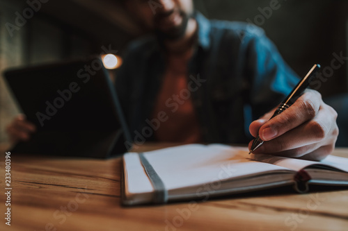 Outgoing man writing in notebook with modern pen while holding digital device in hand. He situating at desk indoor