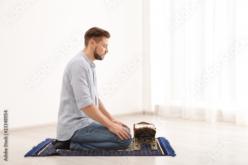 Muslim man with Koran praying on rug indoors. Space for text