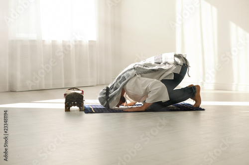 Muslim man in traditional clothes praying on rug indoors