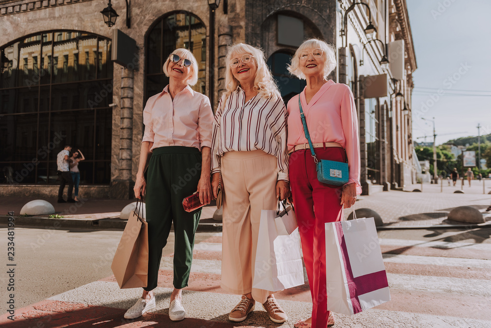Fototapeta premium Full-length photo of three laughing older women holding shopping bags while standing on pedestrian crossing