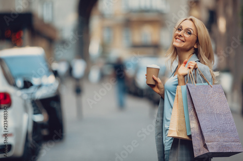 Waist up portrait of charming middle-aged lady holding cup of coffee and shopping bags. She is looking at camera and smiling
