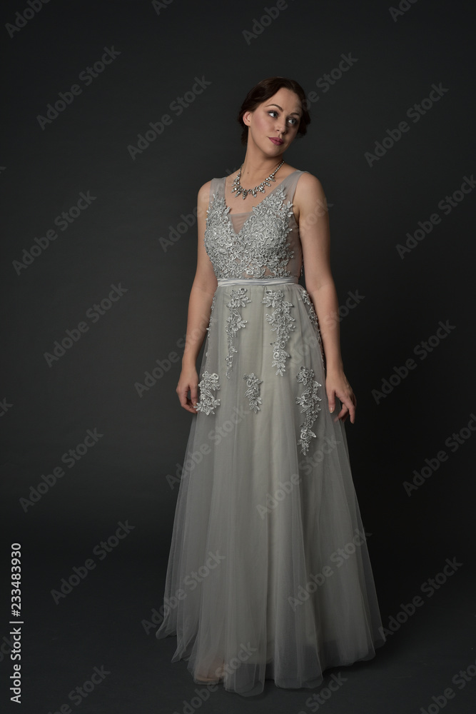 full length portrait of brunette  girl wearing long silver ball gown. standing pose on grey studio background.