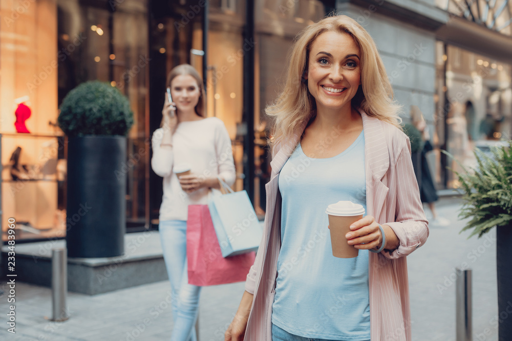 Waist up portrait of beautiful middle-aged woman holding cup of coffee and smiling. Girl with shopping bags having phone conversation on blurred background